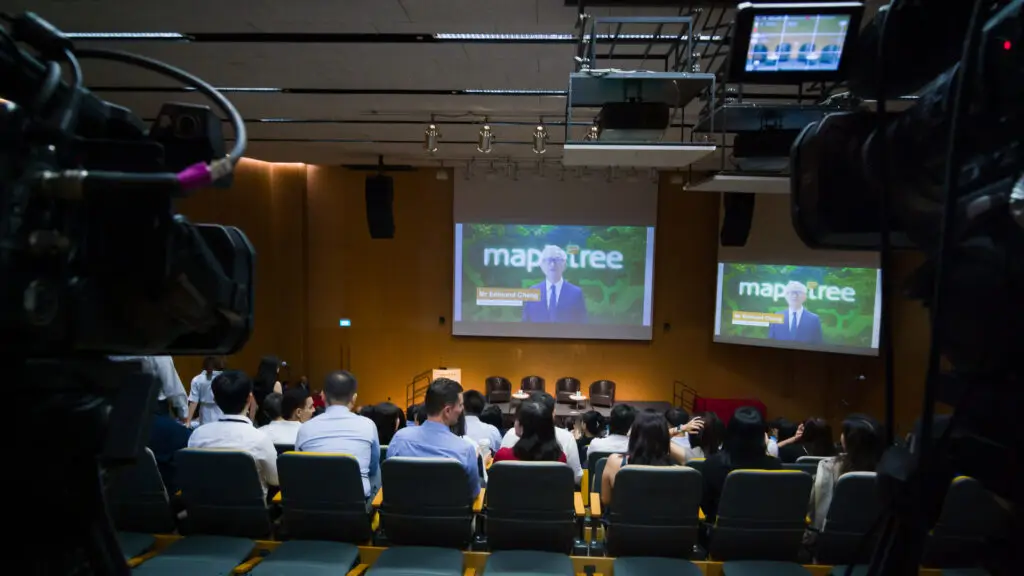 Audience seated in an auditorium during a Mapletree hybrid townhall, with broadcast cameras capturing dual-screen projection for the webcast.