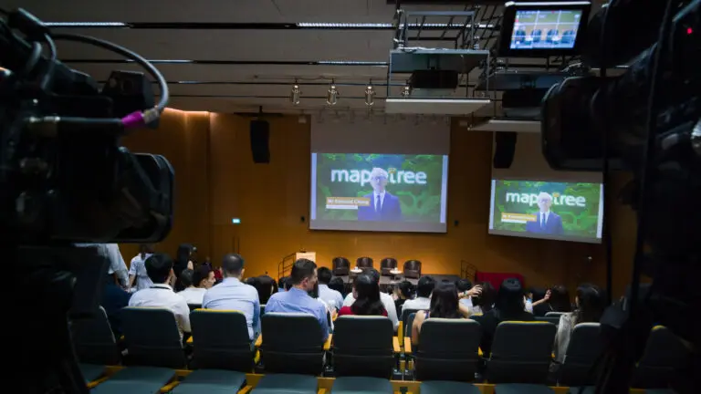 Audience seated in an auditorium during a Mapletree hybrid townhall, with broadcast cameras capturing dual-screen projection for the webcast.